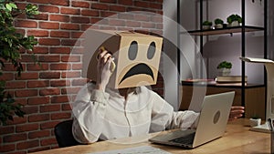 Portrait of a woman in a cardboard box with a negative emoji on her head. Employee sitting at desk, working on laptop