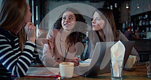 Business lunch concept. Three happy beautiful excited diverse female friends use laptop cheerfully talking at loft cafe.