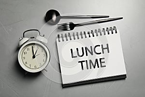 Business lunch. Notebook with phrase Lunch Time, cutlery and alarm clock on light gray table, flat lay