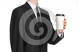 Business lunches coffee theme: businessman in a black suit holding a white blank paper cup of coffee with a brown plastic cap isol