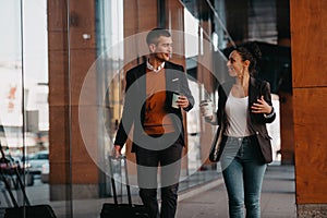 Business man and business woman talking and holding luggage traveling on a business trip, carrying fresh coffee in their