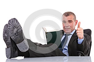 Business man with feet on desk holds a book and shows thumb up