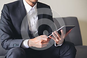 A business man in a gray suit sitting on sofa holding , using and looking at tablet pc for work