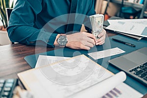 Business man hand writing and signing white blank bank check book and dollar bill, coin, laptop and graph chart on the desk at