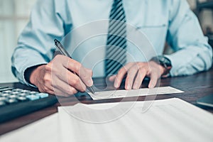 Business man hand writing and signing white blank bank check book and dollar bill, coin, laptop and graph chart on the desk at