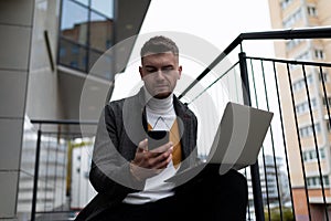 a business man with a laptop and a phone in his hands sits on the stairs next to the office center