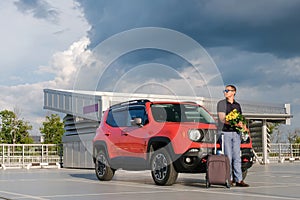 Business man with luggage and flowers stands near his car in the parking lot