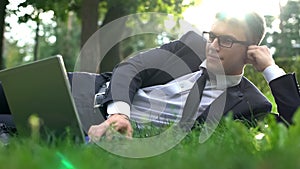 Business man lying on grass in park and working on laptop, harmony with nature