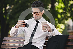 Business man outdoors in the park play games by phone drinking coffee.