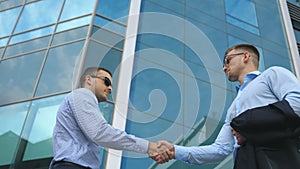 Business man passing a black briefcase to his partner. Colleagues shake hands outdoor in the city background. Two young
