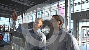 Business man pointing something to his colleague in sunglasses at timetable board screen. Two young businessmen looking