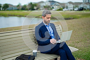 Business man sitting on a bench in park. Man is working on computer outside. Handsome business man making online work