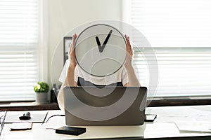 Business man sitting at  desk holds a clock, covering his face