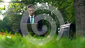 Business man sitting on grass and working on laptop in park outside office