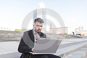 Business man sitting on the street, working on a laptop and looking into the camera