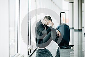 Business man sitting at the terminal airport on the floor with suitcase flight delay, two hands touch at head, headache, waiting t