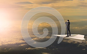 business man standing and spying binocular on papet plane against sun rising over cloud scape