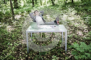 Business man in suit relax at his working office desk in green park with hands over head and legs on table. Great job done. Busine