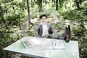 Business man talk on mobile phone at office table in green forest with legs on table. Business concept.