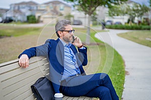 Business man talk on phone sitting on a bench in park. Man in suit call phone outside. Handsome business man using phone