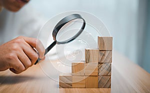 A business man uses a magnifying glass to look at the stacked wooden dice to find profits and investments.