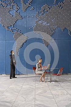 Business man using mobile phone business woman using laptop in office with world map on wall