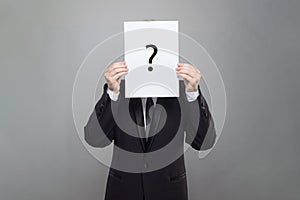 Business man wearing black suit hiding his face behind white paper with black question mark on grey background