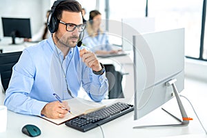 Business man wearing headset looking at computer and taking notes to notebook accompanied with colleagues in background.