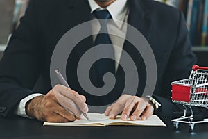 Business man wearing suit and calculates the budget. Shopping cart a on table with note book. Budget of poor low income family.