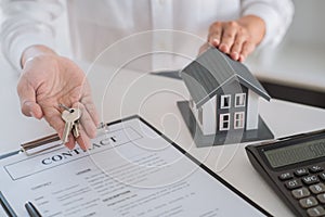 Business. A man wearing white shirt holding the keys the house model  in hands with a house purchase contract below the hands