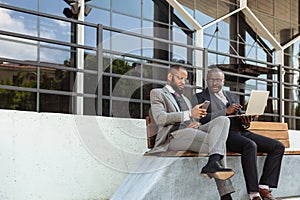 Business meeting of friends outdoors. Two dark-skinned men in suits are sitting on a bench near a city building with a