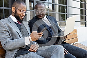 Business meeting of friends outdoors. Two dark-skinned men in suits are sitting on a bench near a city building with a