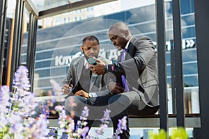 Business meeting of friends outdoors. Two dark-skinned men in suits are sitting on a bench near a city building with a