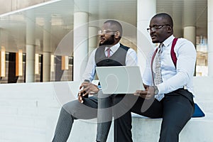 Business meeting outdoors. Two dark-skinned men in suits sit on the steps of a city building with a laptop and have a