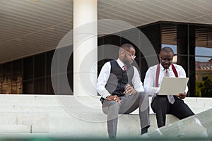 Business meeting outdoors. Two dark-skinned men in suits sit on the steps of a city building with a laptop and have a