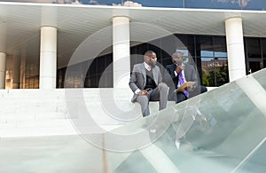 Business meeting outdoors. Two dark-skinned men in suits sit on the steps of a city building with a notebook and a