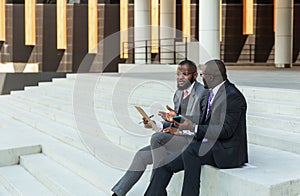 Business meeting outdoors. Two dark-skinned men in suits sit on the steps of a city building with a notebook and a