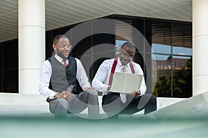 Business meeting of partners outdoors. Two dark-skinned men in suits sit on the steps of a city building with a notebook