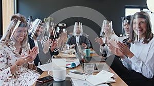 Business meetings during COVID-19. Happy diverse business team listening, clapping at camera, all wearing face shields.