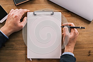 Business mockup, layout. Top view of clipboard with white sheet of paper vertically placed on wooden table. Male hands