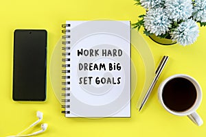 Business Monday motivational words - Work hard, dream big, set goals. With text on book, ear phone, coffee cup, pen and flowers.