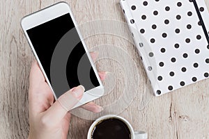 Business and office concept - woman hand holding white smartwhone next to the black and white polka dot cover notebook and cup of