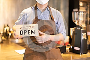 Business owner attractive young Asian man in apron hanging we`re open sign on front door  welcoming clients to new cafe. Happy