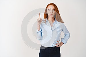 Business people. Attractive female manager with ginger hair and blue eyes pucker lips, raising finger and pointing up