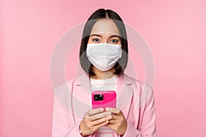 Business people and covid-19 concept. Japanese corporate office lady in suit and medical face mask, using mobile phone