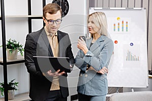 Business people with laptop discussing new projects in office. A young woman and a man stand near flipcharts and solve work issues