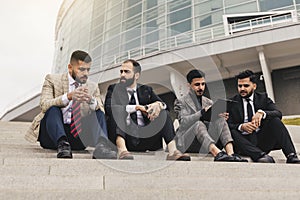 Business people outdoor meeting. A company of male businessmen in suits are sitting on the steps of the stairs. Working