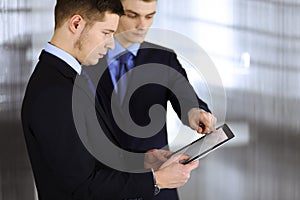 Business people, using a tablet computer, while standing in a modern office. Unknown businessman with a colleague at