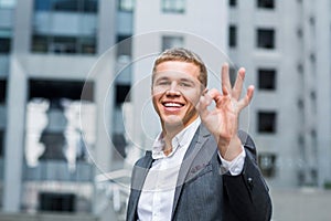 Business, people, vision and success concept - happy smiling businessman in eyeglasses and suit showing ok sign over office buildi