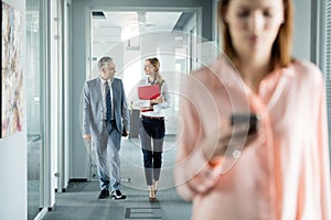 Business people walking in office corridor with female colleague using mobile phone in foreground